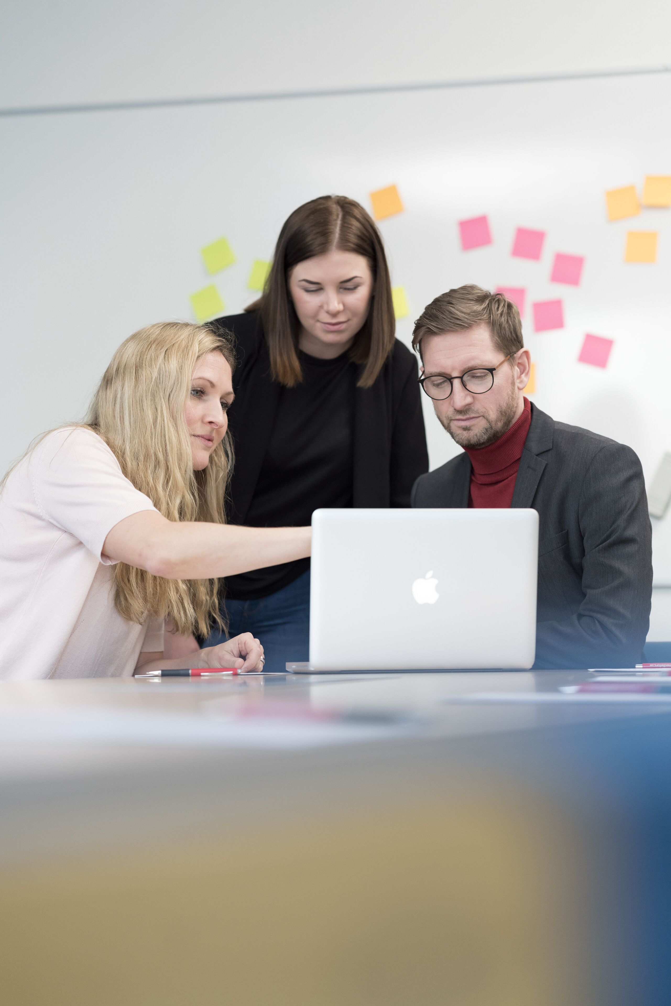 Trois personnes sont réunies autour d’un ordinateur portable dans une pièce où se trouvent un tableau blanc et des post-it.