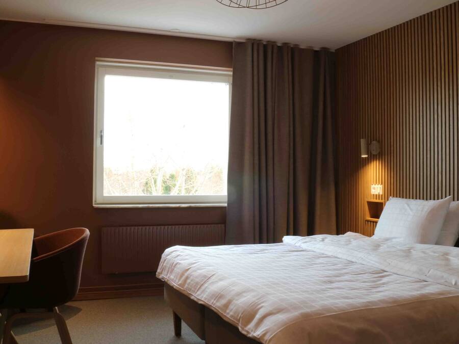 A hotel room with a bed, a desk, a chair, a window, and a wall-mounted lamp.