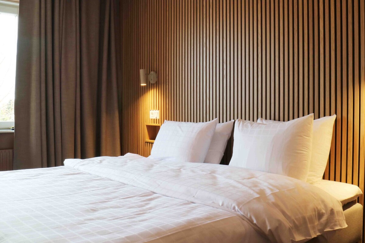 Une chambre d’hôtel avec un lit, des rideaux, un mur recouvert de lambris en bois verticaux et une table de chevet avec une lampe.