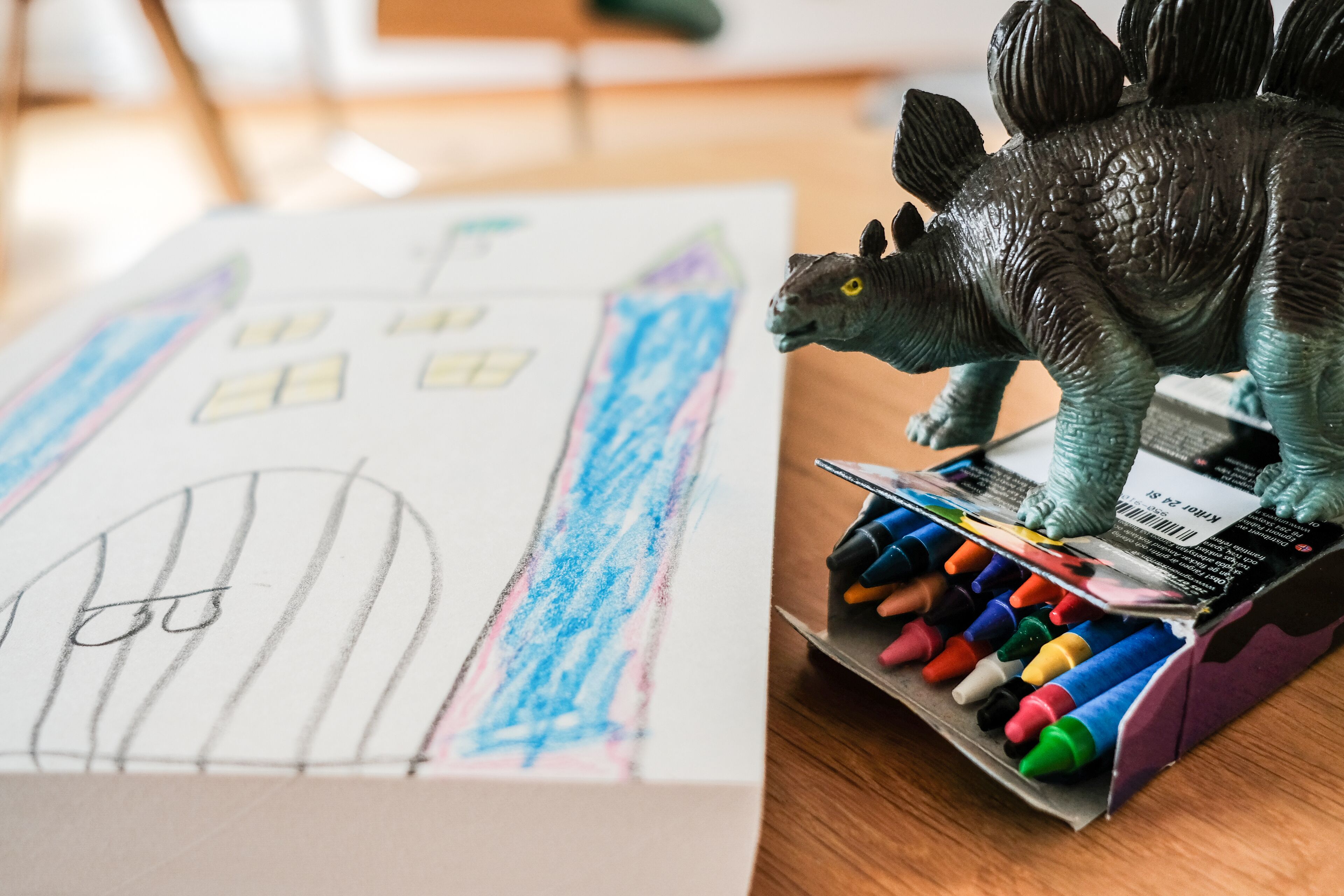 En dinosaurieleksak ligger på en ask med kritor bredvid en teckning av ett hus.