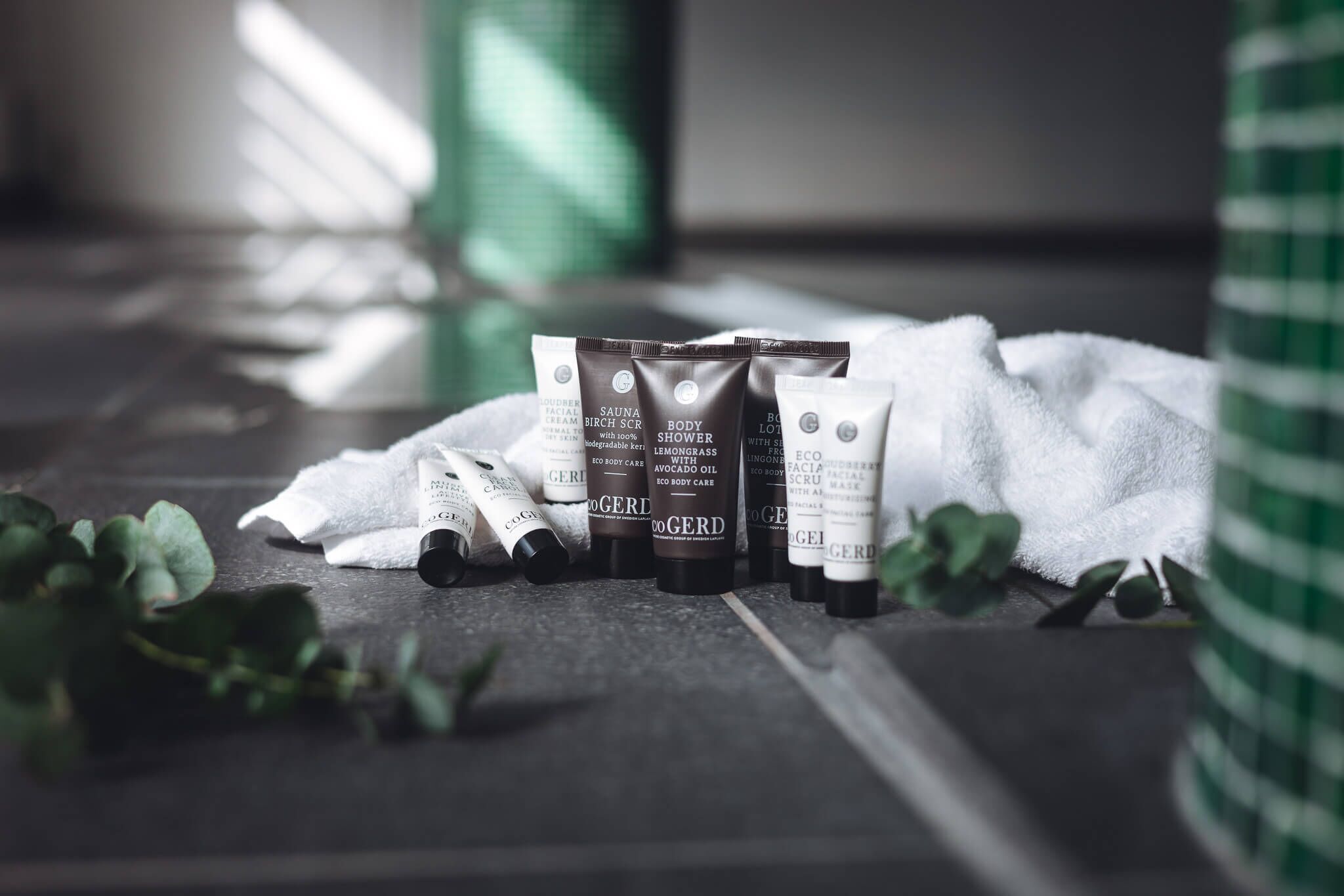 A set of body care products on a dark surface with a white towel and green leaves.