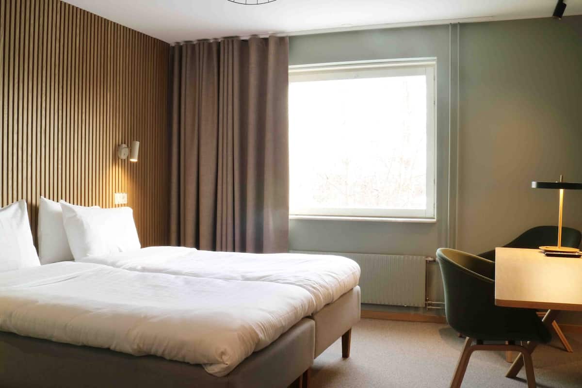 Ein Hotelzimmer mit einem Bett, einem Schreibtisch, einem Sessel, einem Fenster mit Vorhängen und einem an der Wand befestigten Fernseher.