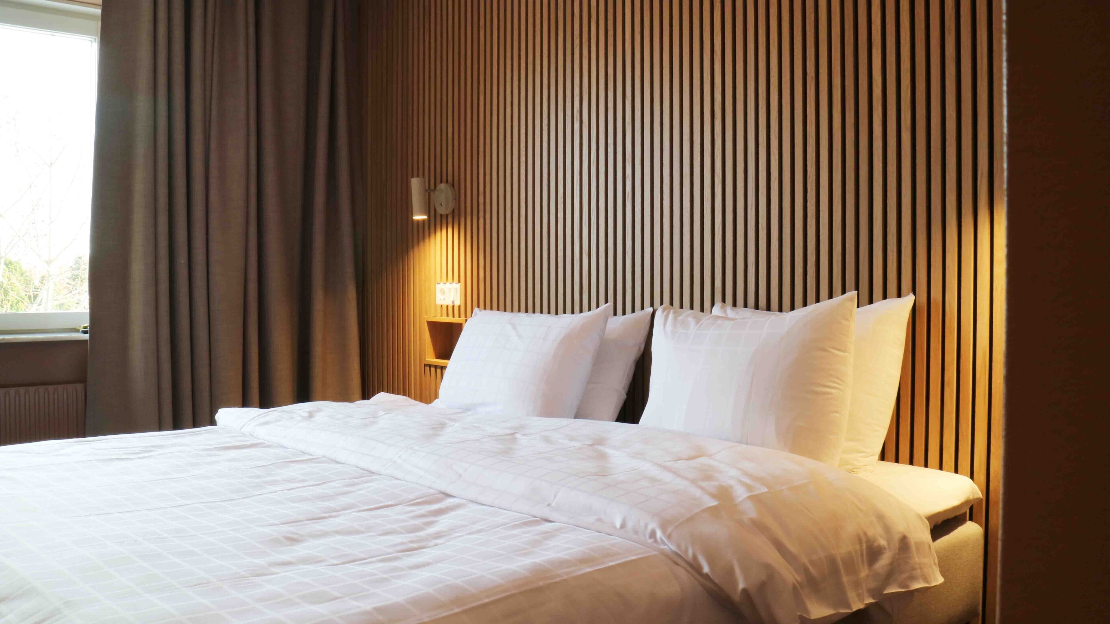 A neatly made bed with white linens in a hotel room with wooden paneling on the walls and a window with curtains.