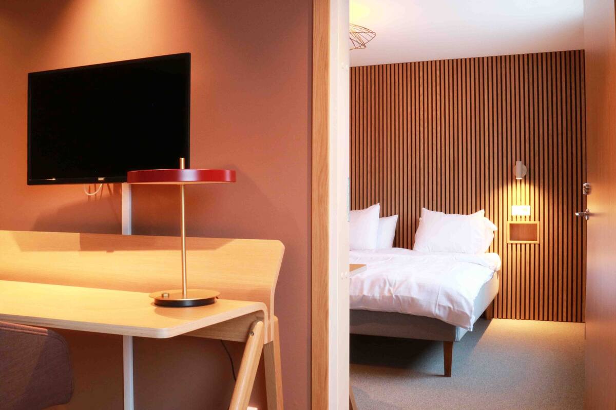 Ein Hotelzimmer mit Bett, Wandfernseher, Schreibtisch mit Stuhl und Nachttisch.
