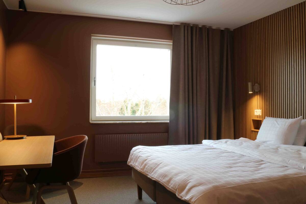 Ein Hotelzimmer mit Bett, Schreibtisch, Stuhl, Fenster und einer Wandleuchte.