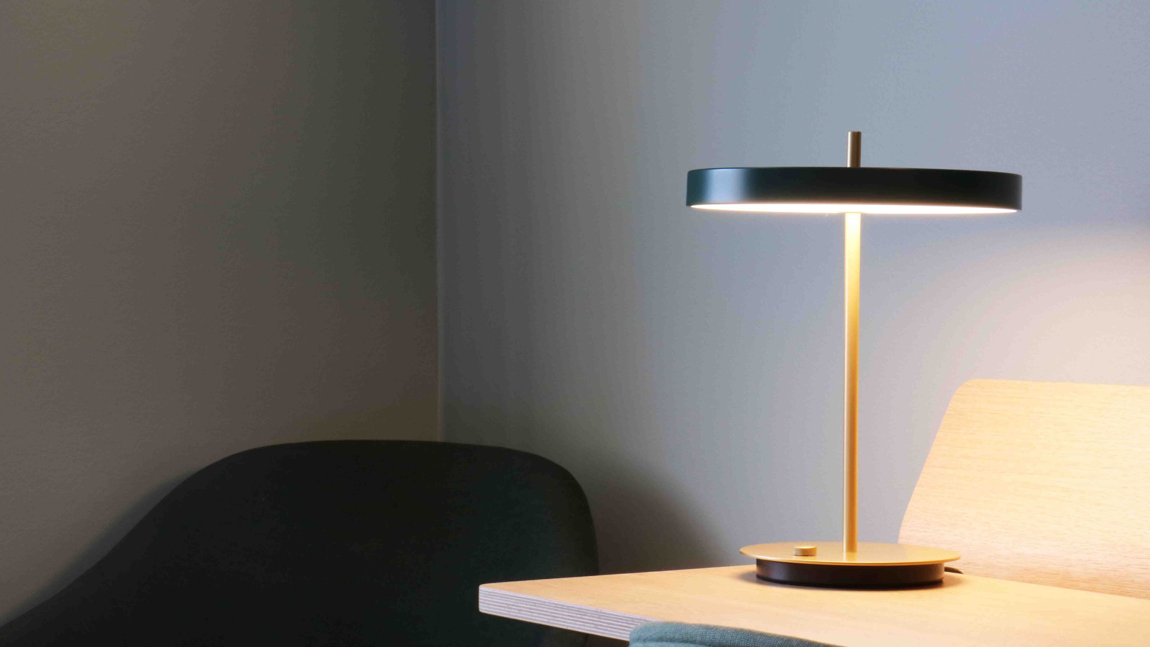 Een moderne bureaulamp met een zwarte kap en een houten voet, geplaatst op een houten tafel tegen een effen muur.