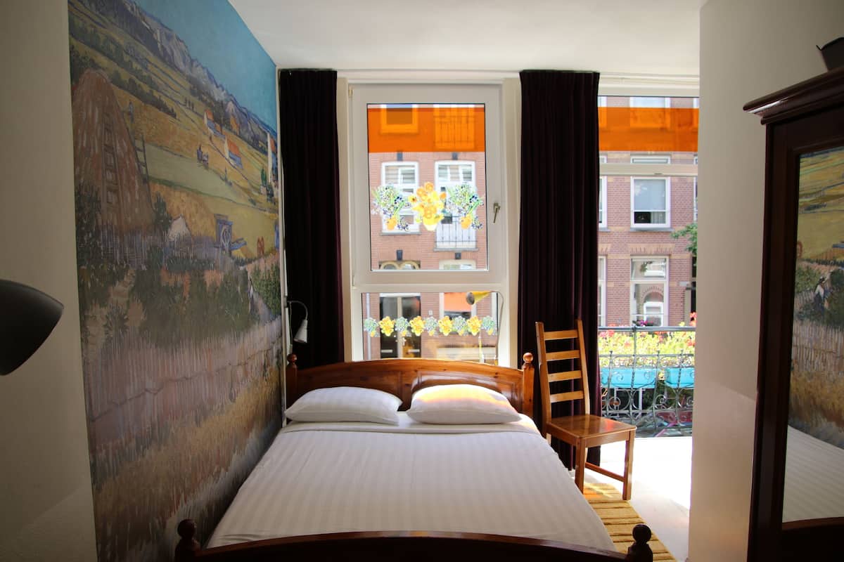 A bedroom with a large bed, a wooden chair, a window with curtains, and a view of buildings outside.