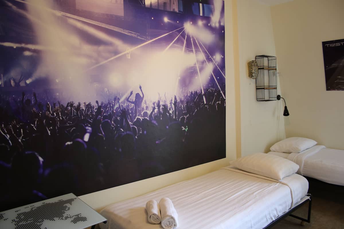 A hotel room with a bed, a wall-mounted light fixture, and a poster of a concert scene.