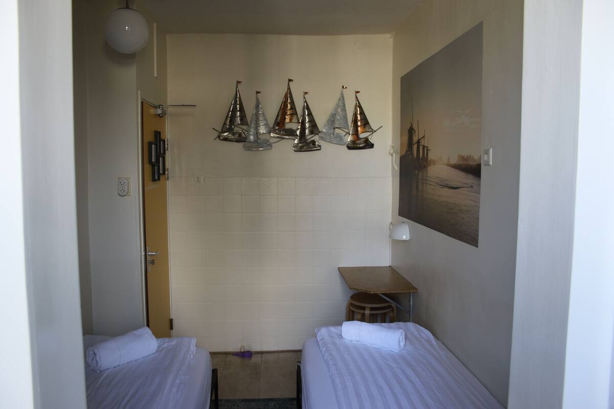 A hotel room with two beds, a small table, a chair, a wall-mounted shelf with decorative items, and a painting of a landscape.