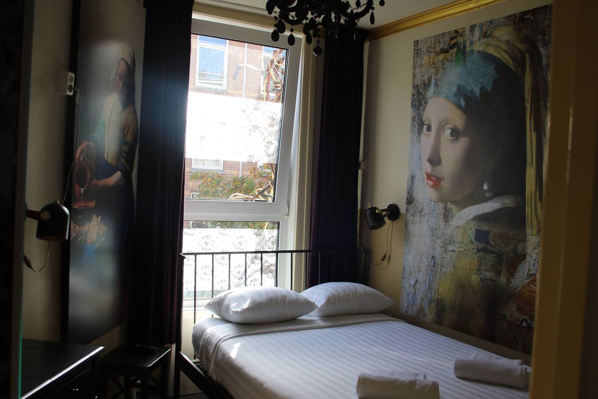 A bedroom with a bed, pillows, a window, and a painting on the wall.
