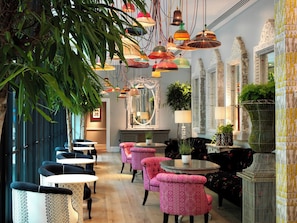 A restaurant interior with round tables, pink upholstered chairs, hanging pendant lights, and a mirror above the bar area. - Ham Yard Hotel, Firmdale Hotels (London)