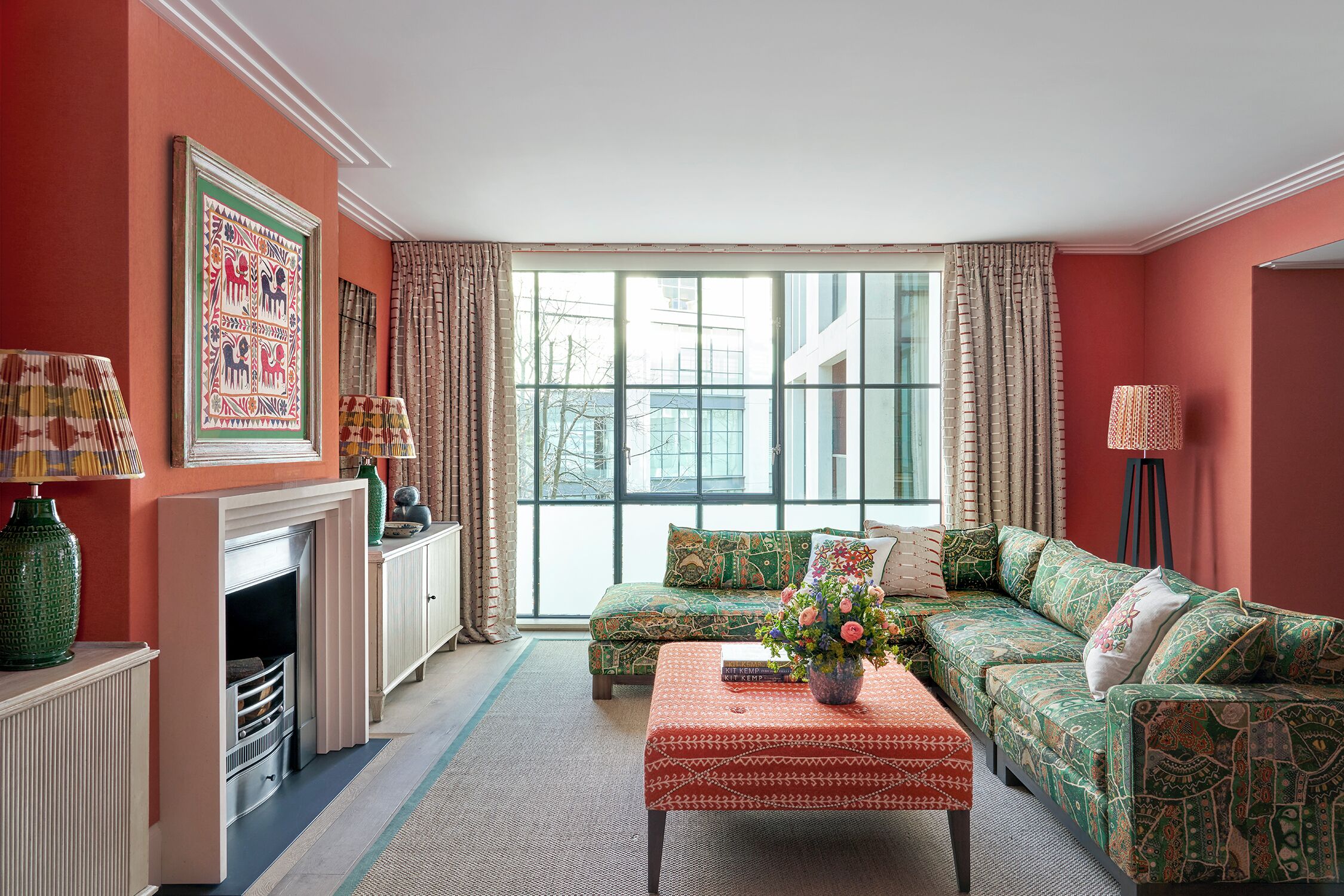 A living room with a fireplace, a sofa set, a coffee table with a vase, and a large window with curtains.