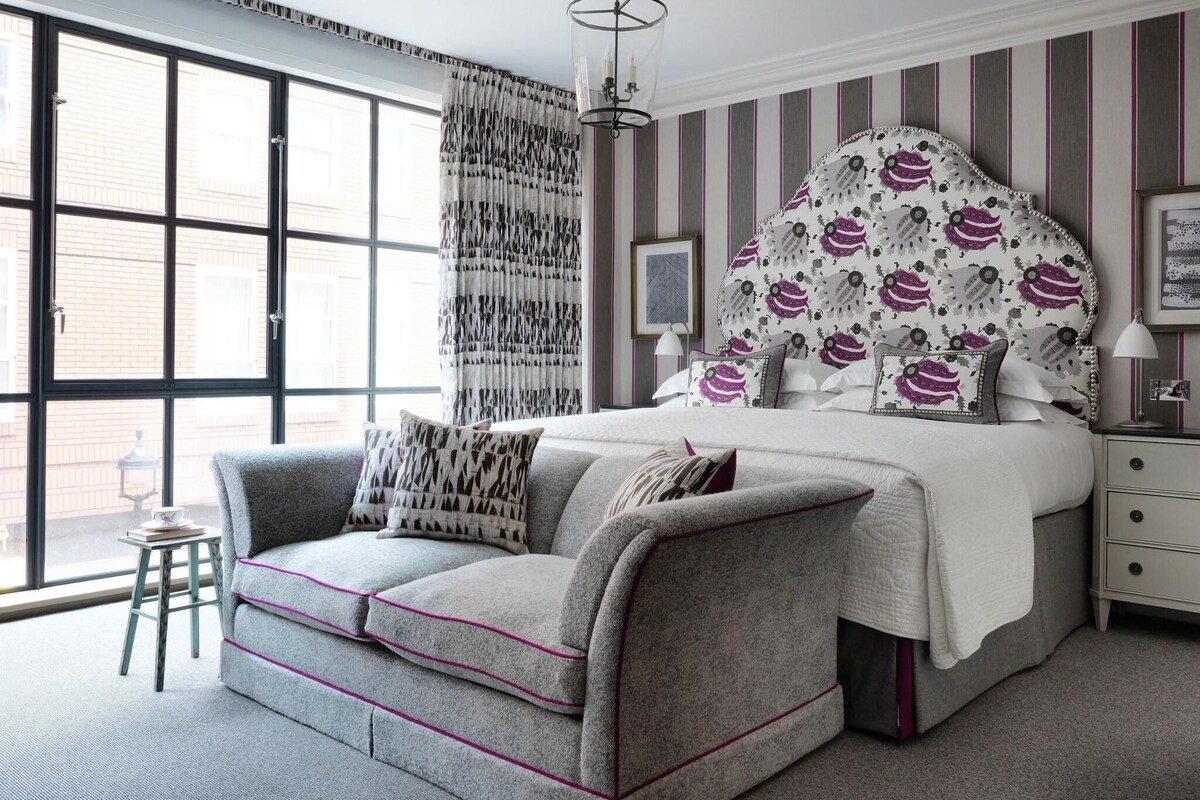 A bedroom with a large window, a grey sofa, a bed with a patterned headboard, and a striped wall.