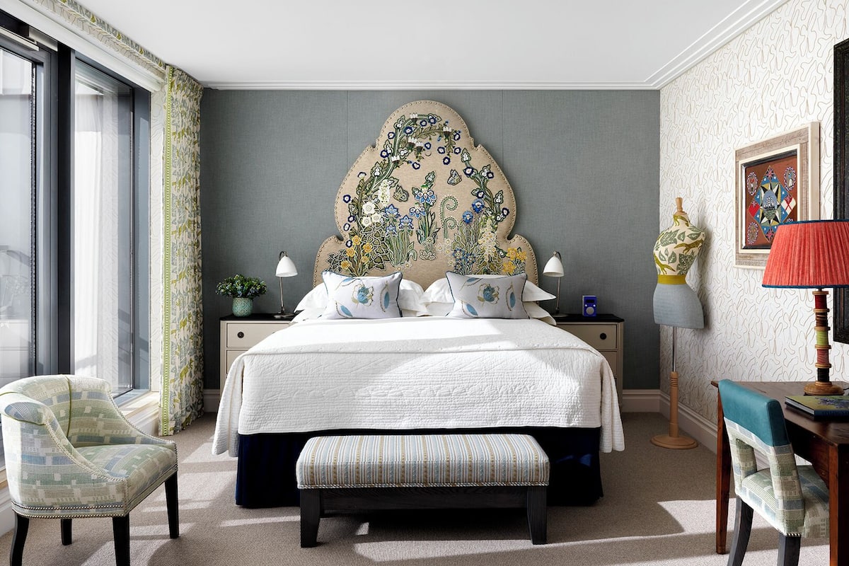 A bedroom with a large bed, a chair, a bench, a nightstand, a lamp, and a window with curtains.