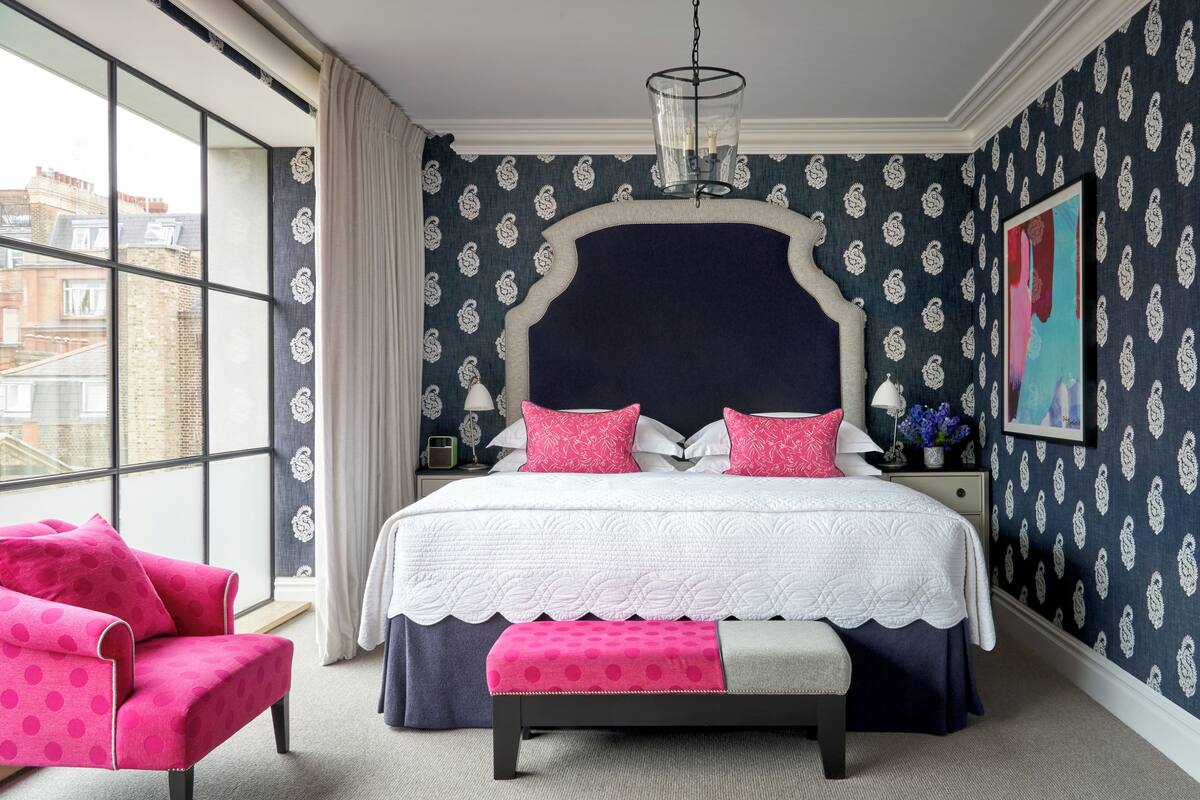 A bedroom with a large window, a bed with a white and navy headboard, pink and navy bedding, a pink armchair, and a bench.