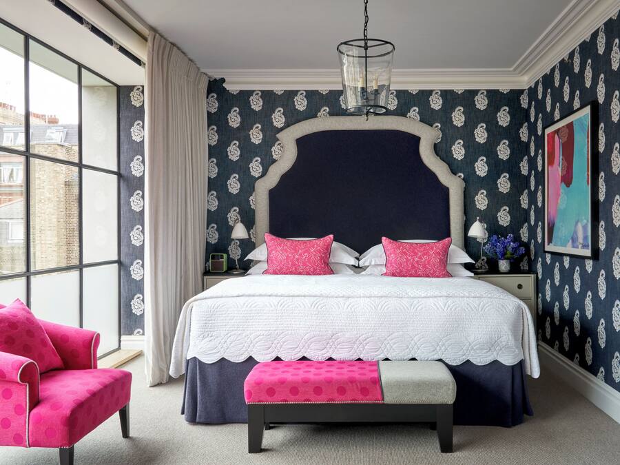 Un dormitorio con un ventanal grande, cama con cabecera blanca y azul marino, ropa de cama rosa y azul marino, un sillón rosa y un banco.