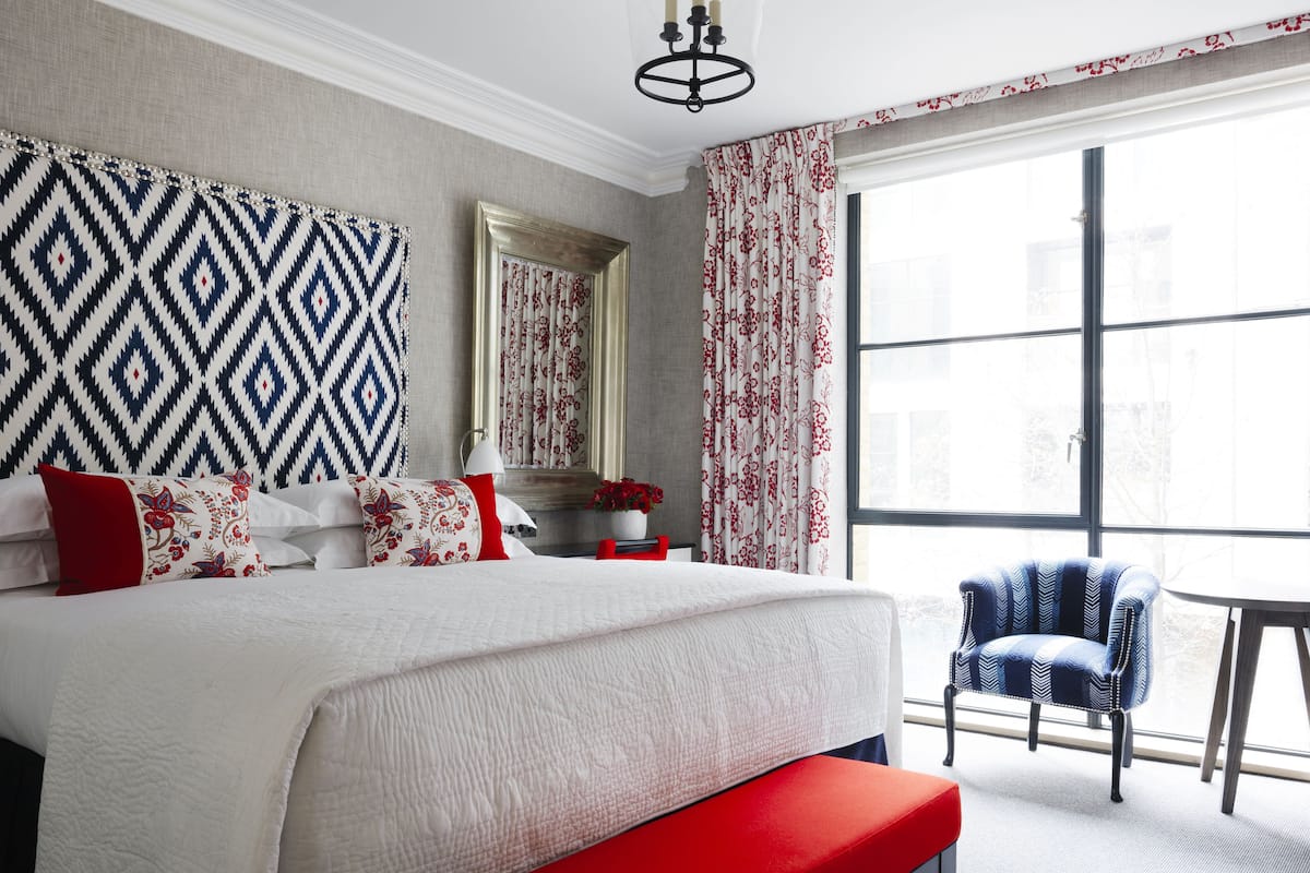 A hotel room with a large bed, a bench, a chair, a mirror, and a window with curtains.