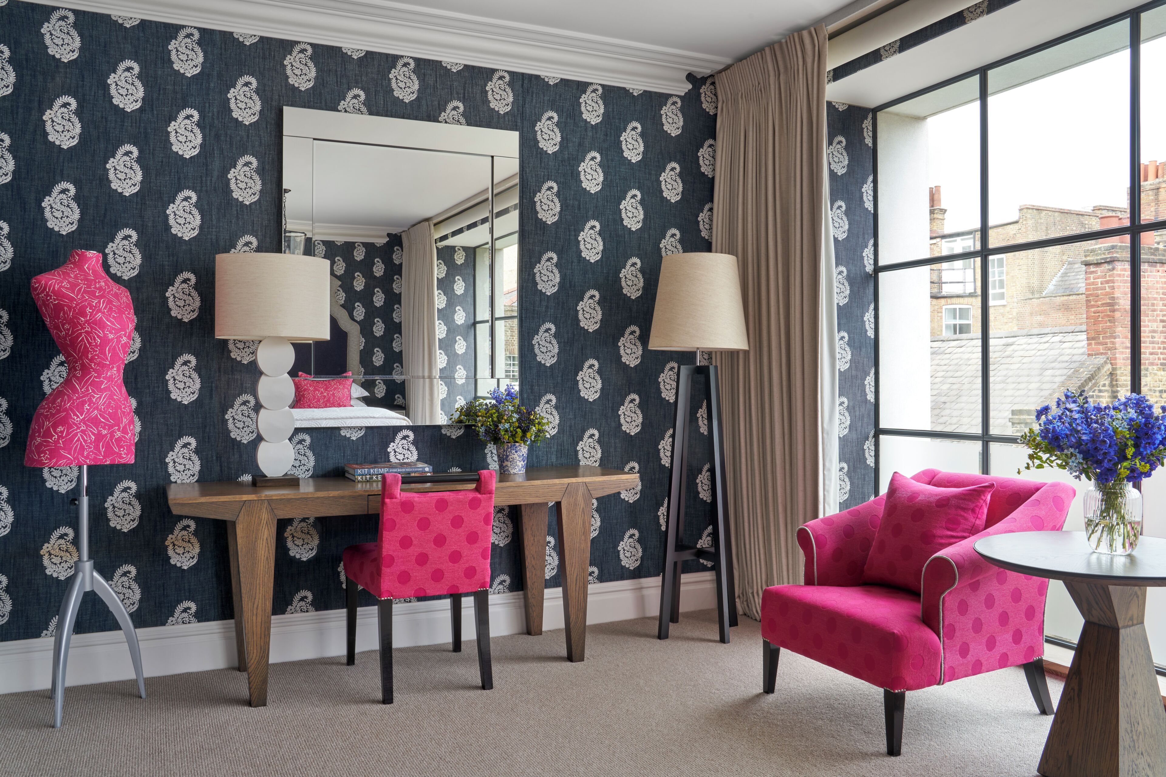 Una habitación con papel tapiz estampado, un escritorio de madera, una silla, un sillón rosa, un espejo y una ventana con vistas a edificios.