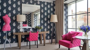 Una habitación con papel tapiz estampado, un escritorio de madera, una silla, un sillón rosa, un espejo y una ventana con vistas a edificios.