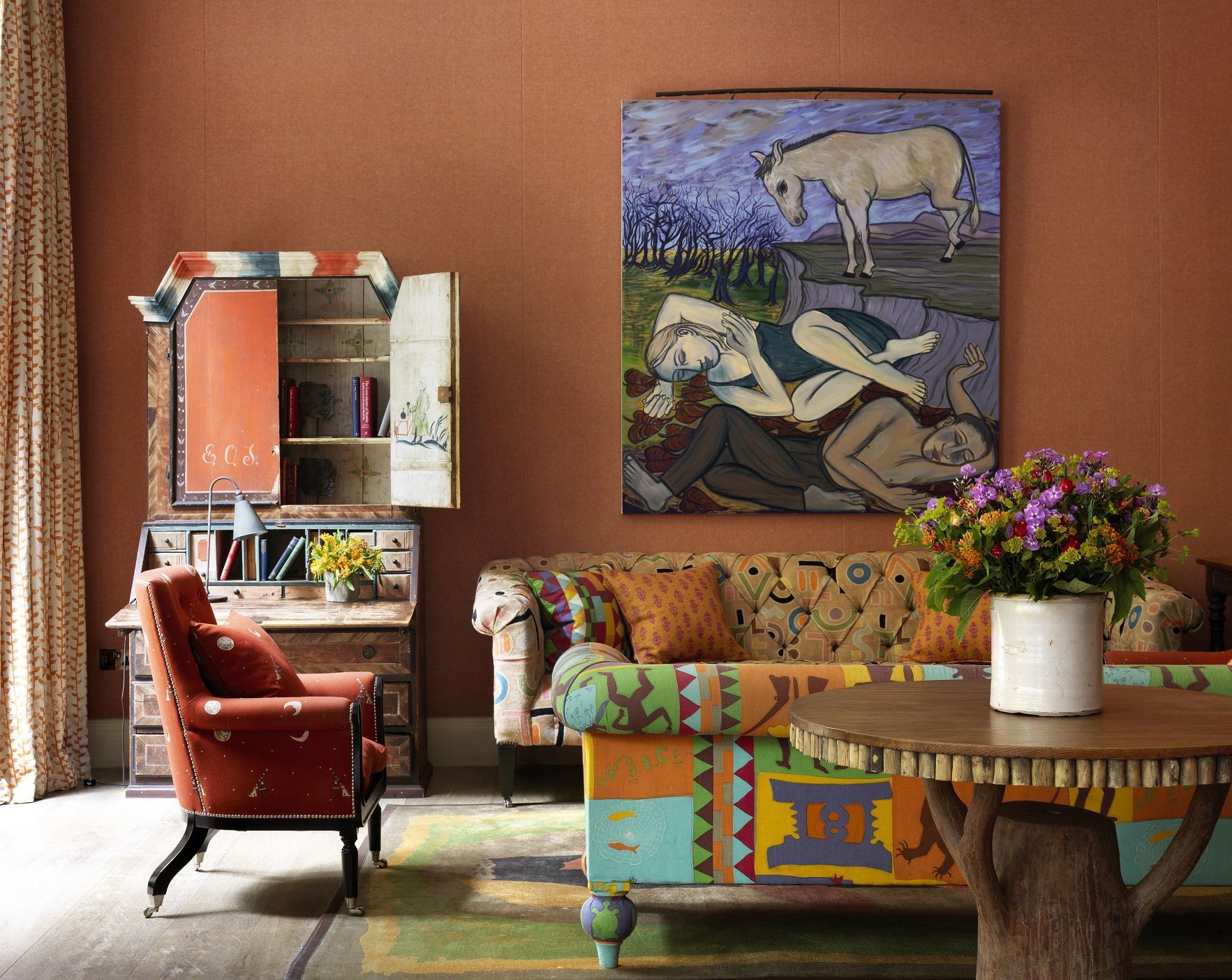Una sala de estar acogedora con un sillón rojo, un sofá colorido, una mesa de centro de madera y un cuadro de un caballo y una mujer en la pared.