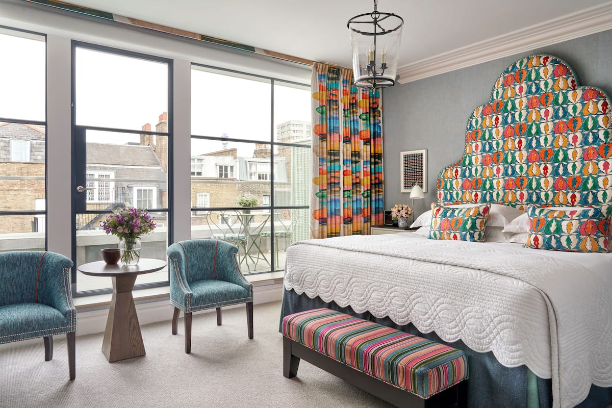 A bedroom with a large window, a bed with a patterned headboard, two chairs, a small table with a vase, and a bench.