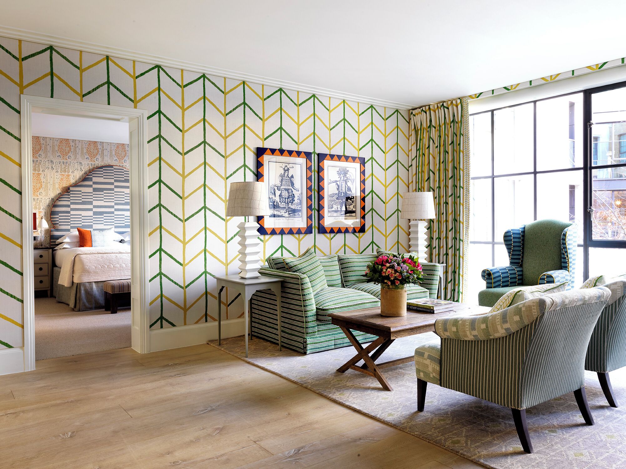 A room with a chevron-patterned wallpaper, a bed with a patterned headboard, a green sofa, a striped armchair, and a table with a vase of flowers.