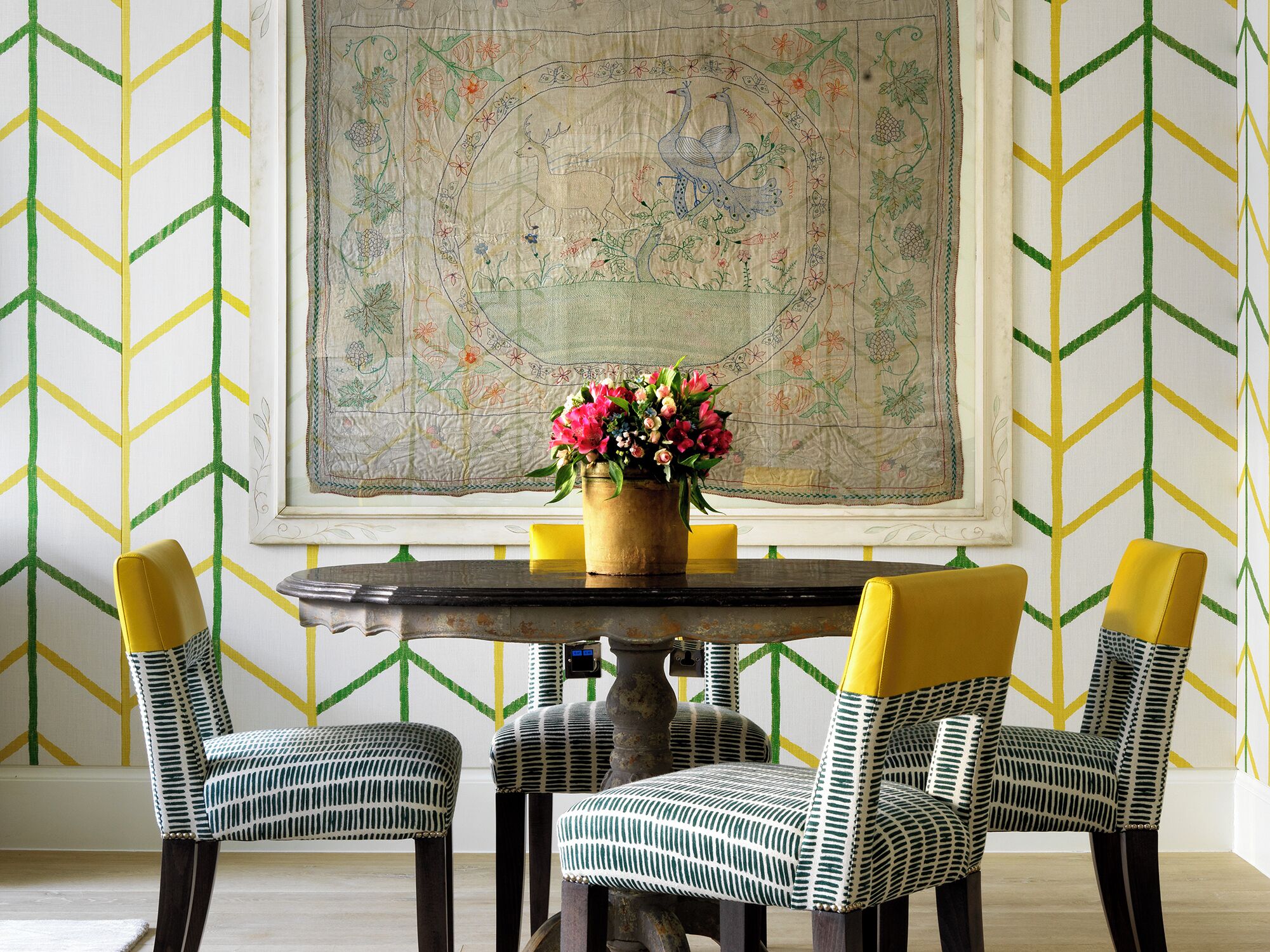 A dining area with a round table, four chairs with yellow backs, and a floral centerpiece.