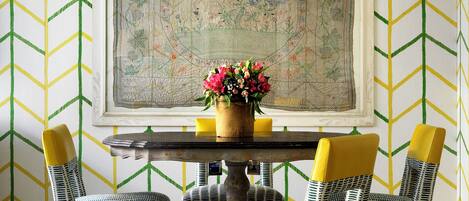 A dining area with a round table, four chairs with yellow backs, and a floral centerpiece.
