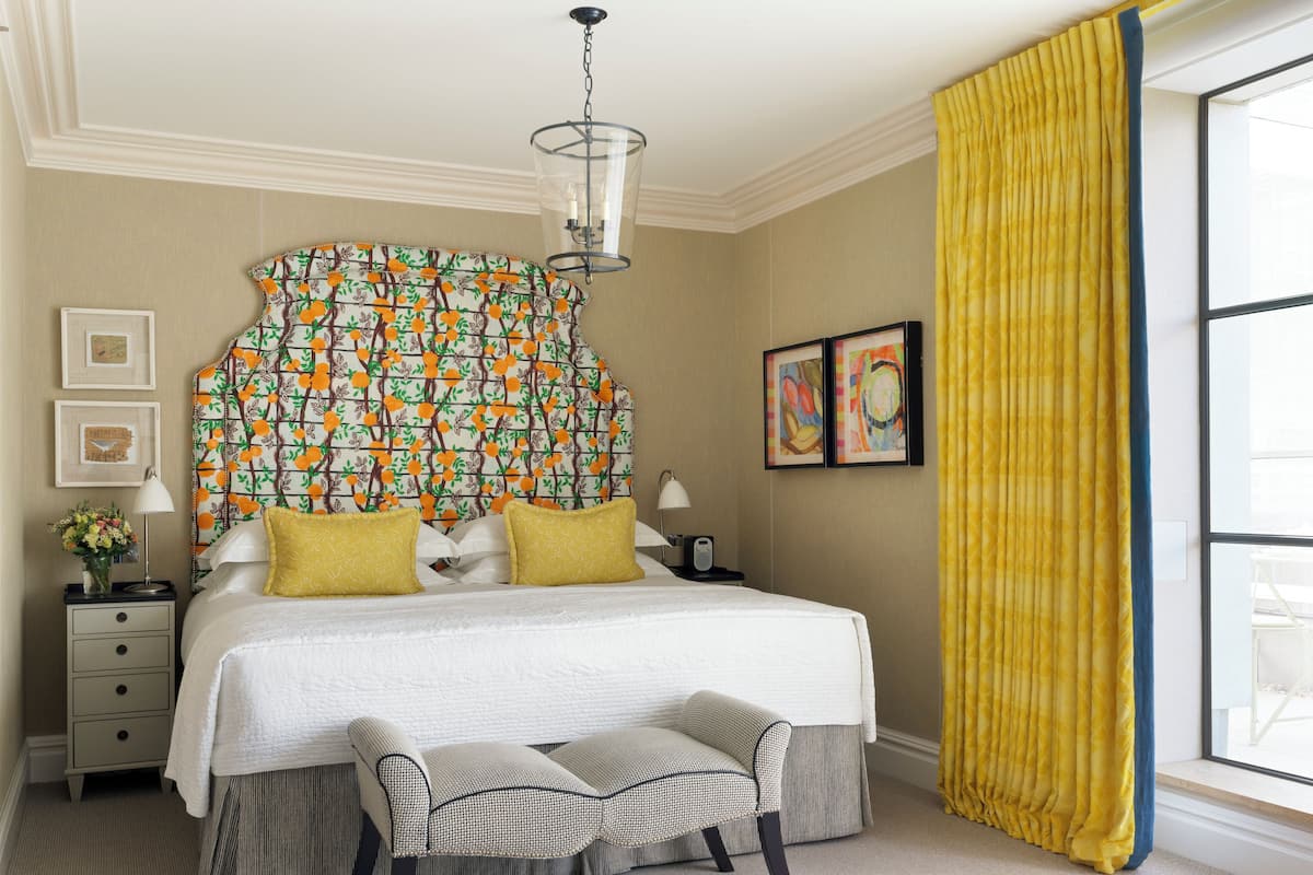 A neatly arranged bedroom with a patterned headboard, a bedside table with a lamp, a bench, and a large window with yellow curtains.