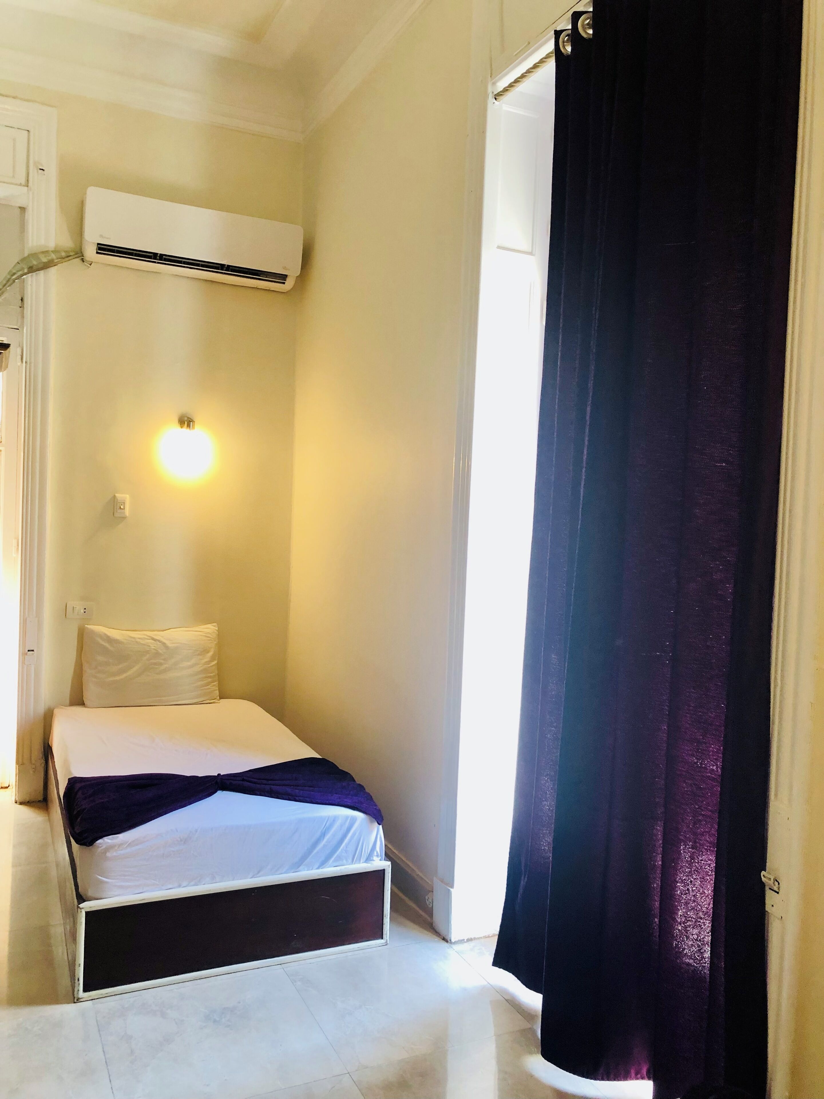 A small, single-bed room with a white mattress, a black headboard, a wall-mounted air conditioner, a bedside lamp, and a window with a black curtain.