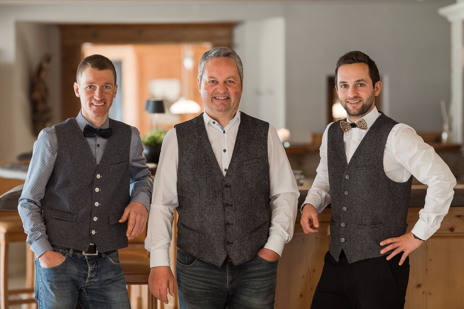Drei Männer in festlicher Kleidung stehen in einem Raum mit Holzmöbeln und modernem Interieur.