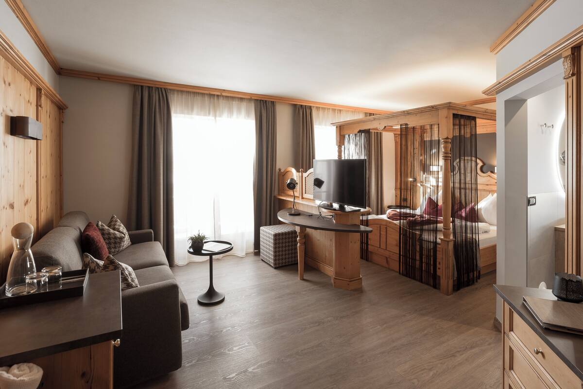 Moderni hotellihuone, jossa on sohva, pieni ruokapöytä, televisio ja sänky, jonka päädyssä on puinen koristepaneeli.