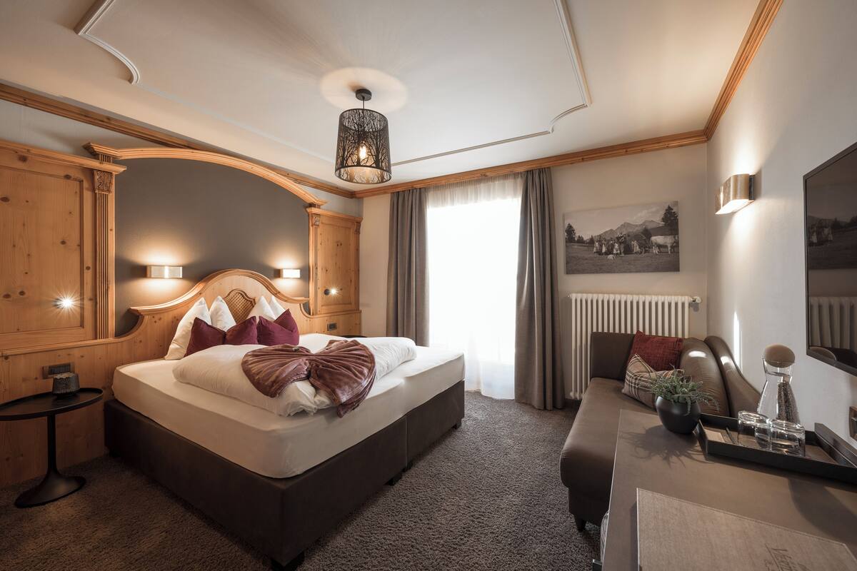 Moderni hotellihuone, jossa on suuri sänky, puinen pääty, pieni sivupöytä, työpöytä, jossa on tarjotinalusta, sekä suuri ikkuna, jossa on verhot.