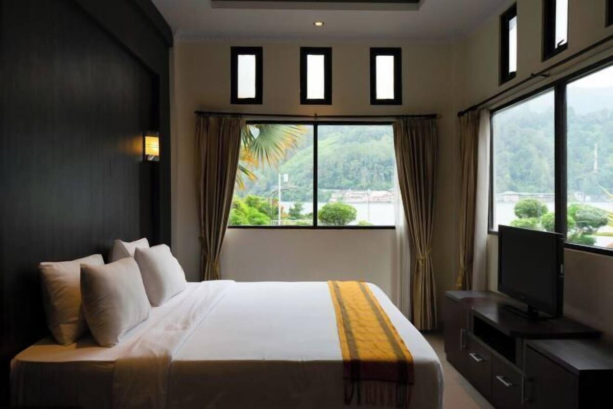 A hotel room with a bed, a television, a window with curtains, and a view of greenery.