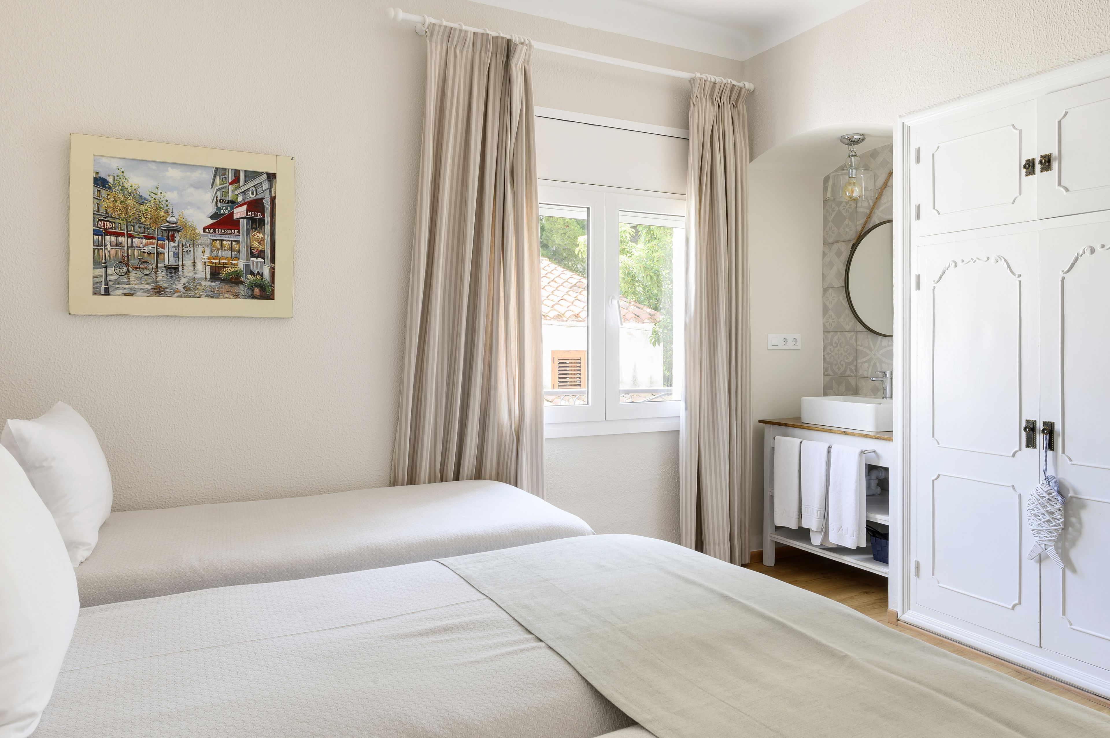 A bedroom with two beds, a window with curtains, a bathroom with a mirror and towel rack, and a painting on the wall.