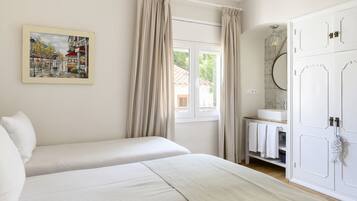 A bedroom with two beds, a window with curtains, a bathroom with a mirror and towel rack, and a painting on the wall.
