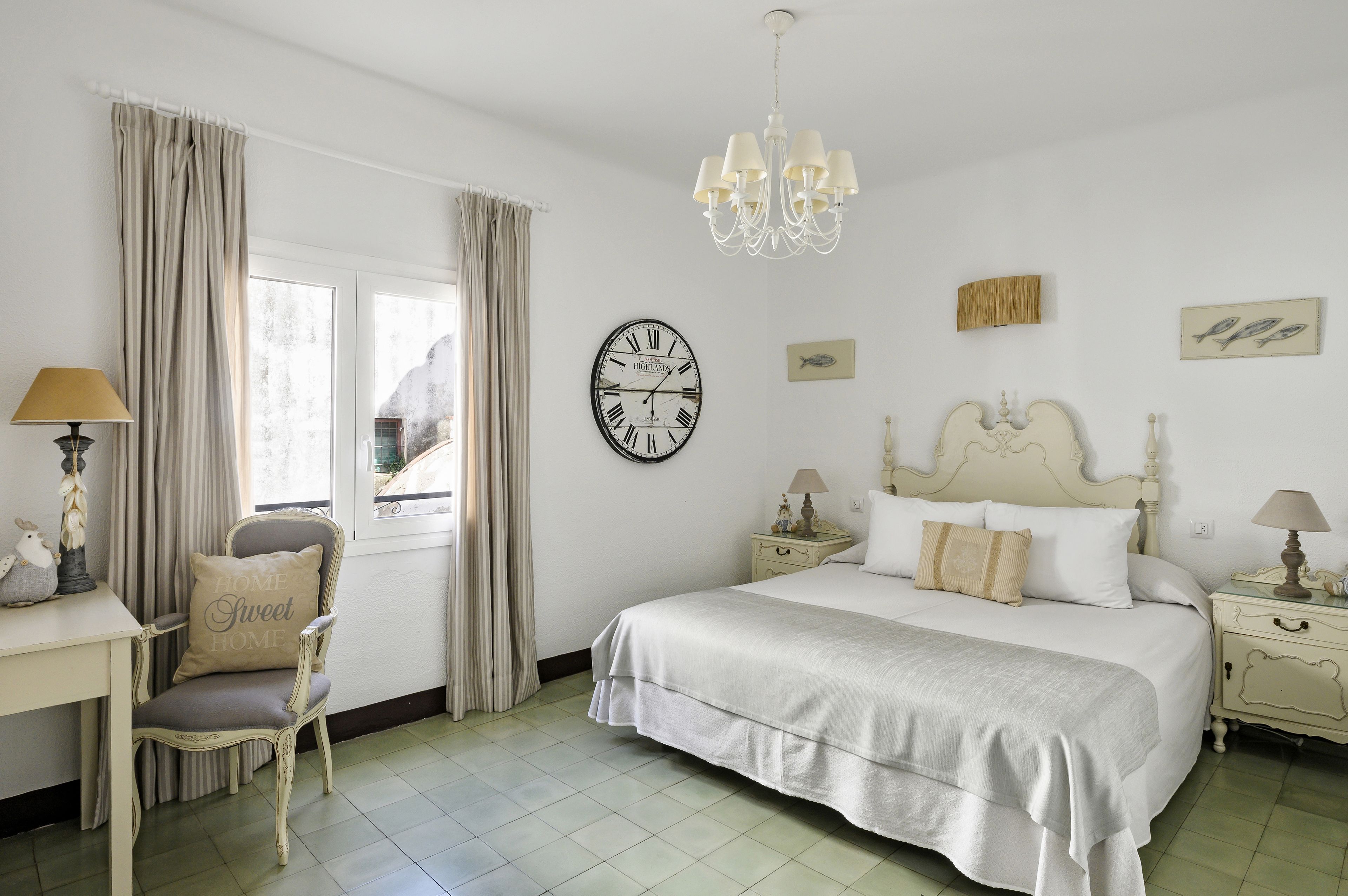 Une chambre à coucher comprenant un lit, une chaise, une table de chevet, une lampe, une horloge et une fenêtre avec des rideaux.