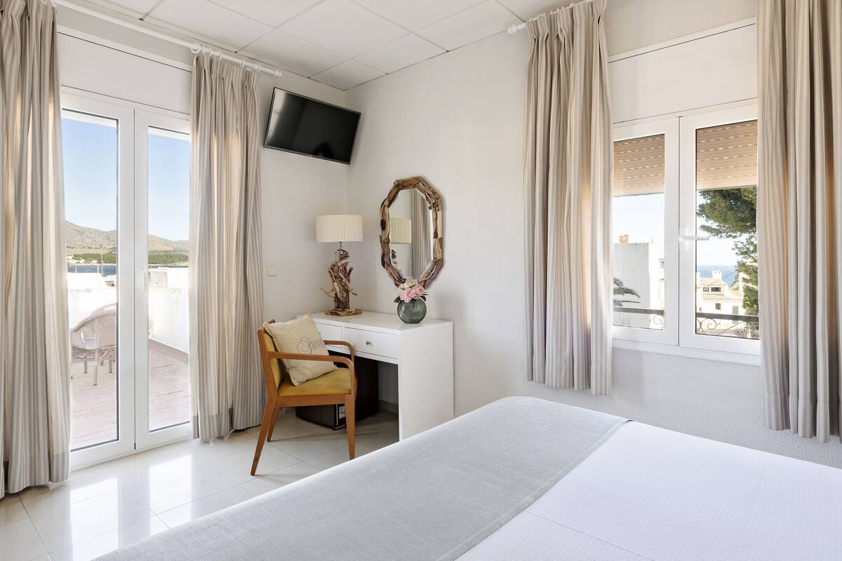 A hotel room with a bed, a desk with a chair, a TV, a mirror, and a view of the outdoors.