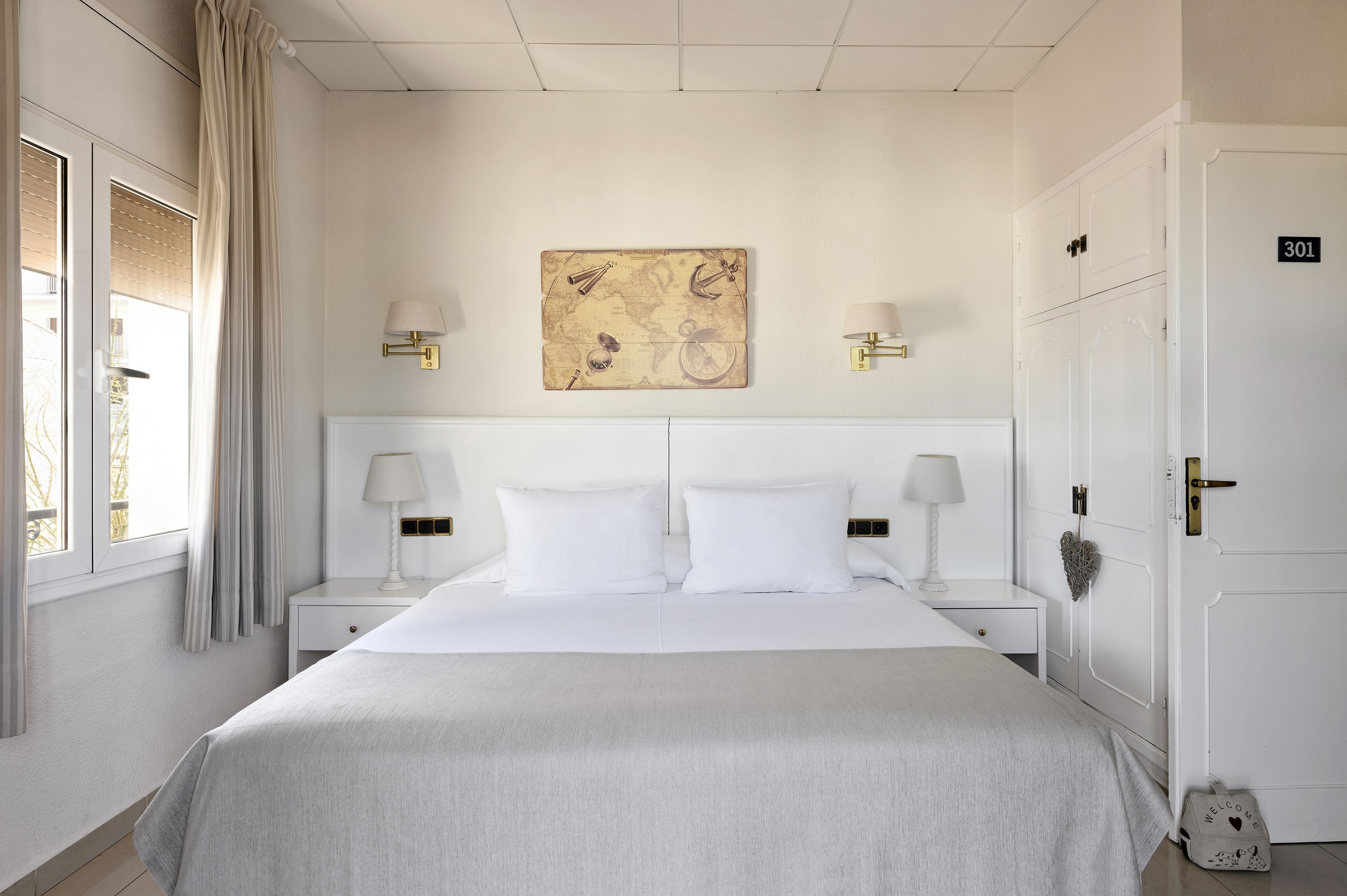 A neatly arranged hotel room with a large bed, two bedside tables with lamps, a map artwork on the wall, and a door with the number 301.