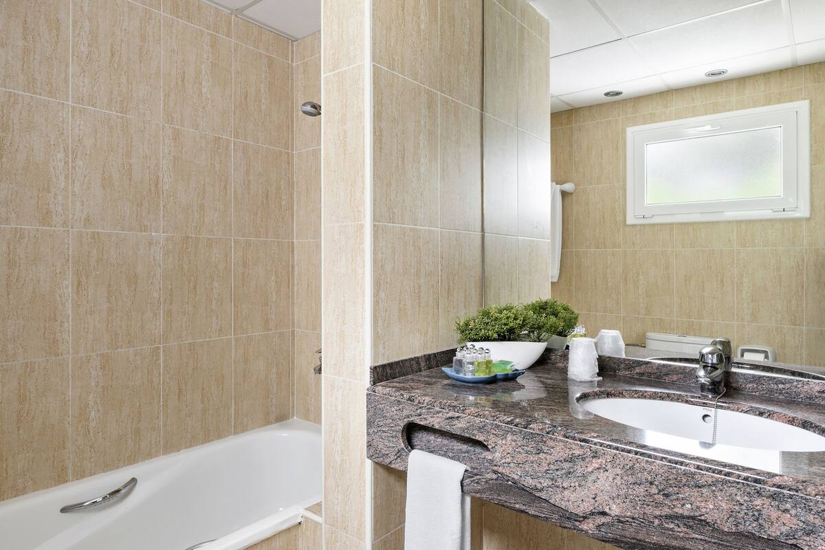 A bathroom with beige tiles, a white bathtub, a granite countertop, a sink, a mirror, and a potted plant.