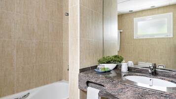 Une salle de bain avec des carreaux de couleur beige, une baignoire blanche, un comptoir en granit, un lavabo, un miroir et une plante en pot.