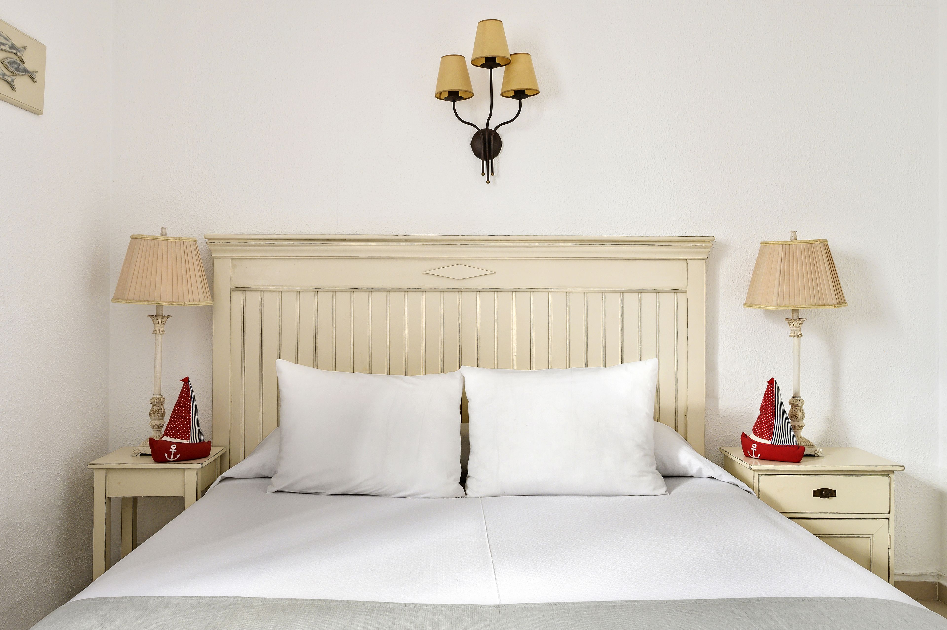 Una cama bien tendida con sábanas blancas, dos mesitas de noche con lámparas y una lámpara fijada a la pared.