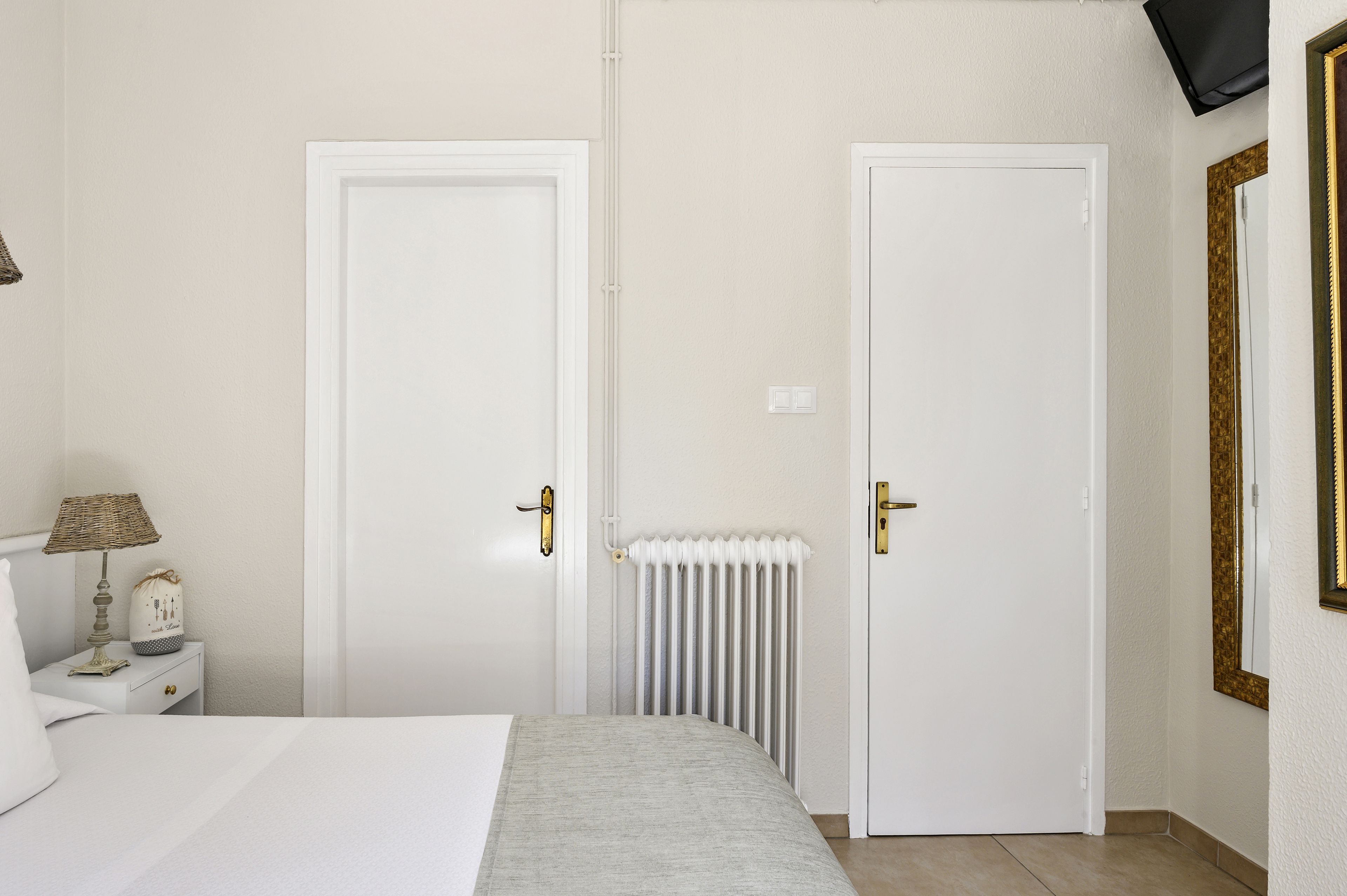Une chambre à coucher avec deux portes blanches, une table de chevet avec une lampe, un radiateur et un lit recouvert d’une couverture grise.