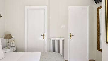 A bedroom with two white doors, a bedside table with a lamp, a radiator, and a bed with a grey bedspread.