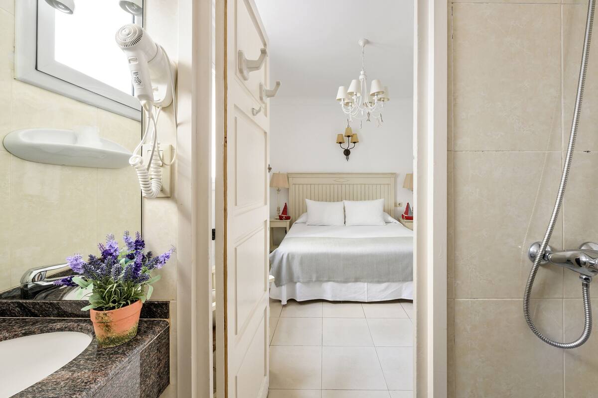 A bathroom with a sink, a hairdryer, a potted plant, a shower, and a bed with white bedding.