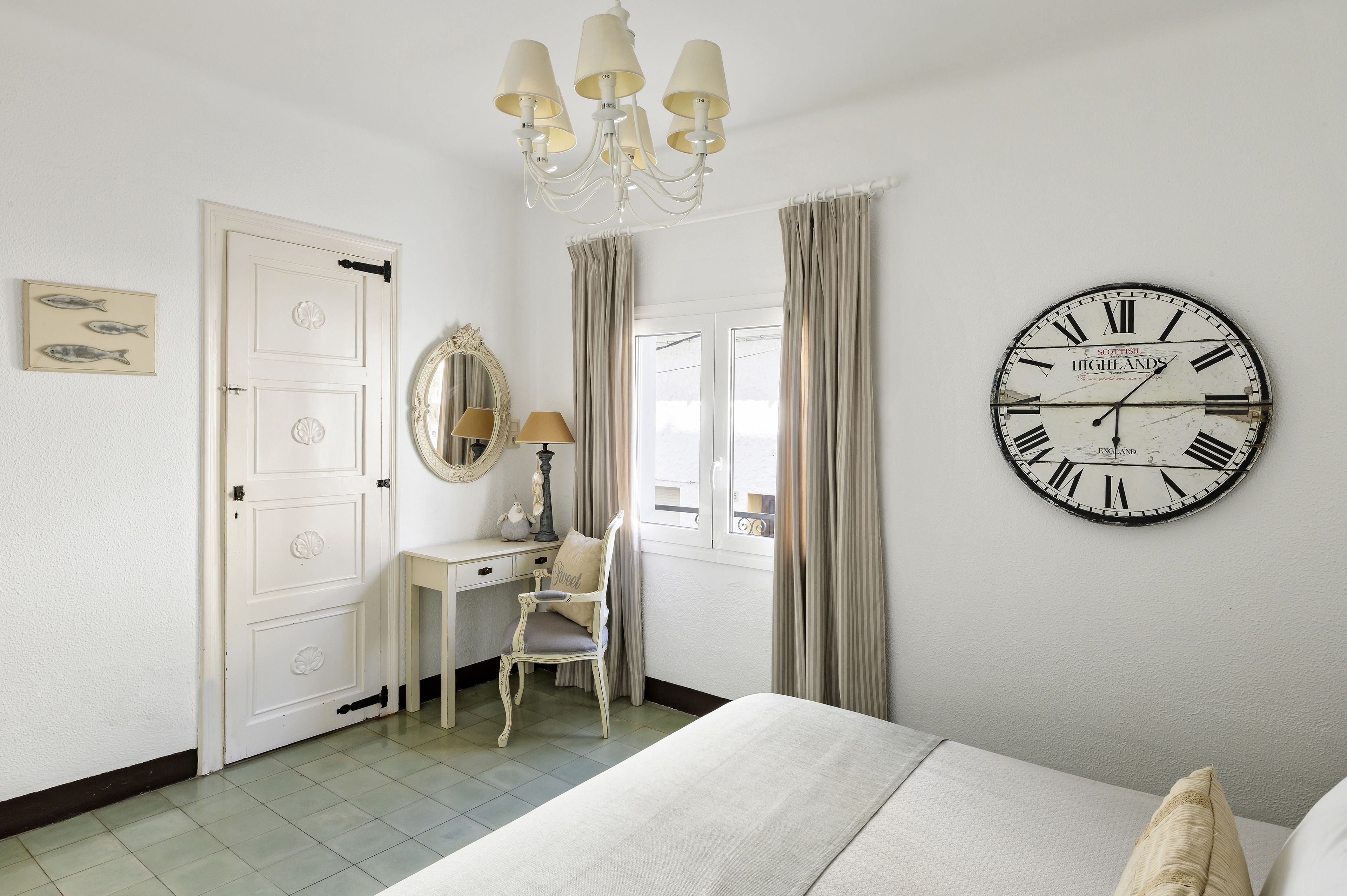 A bedroom with a bed, a desk with a chair, a large wall clock, and a chandelier.