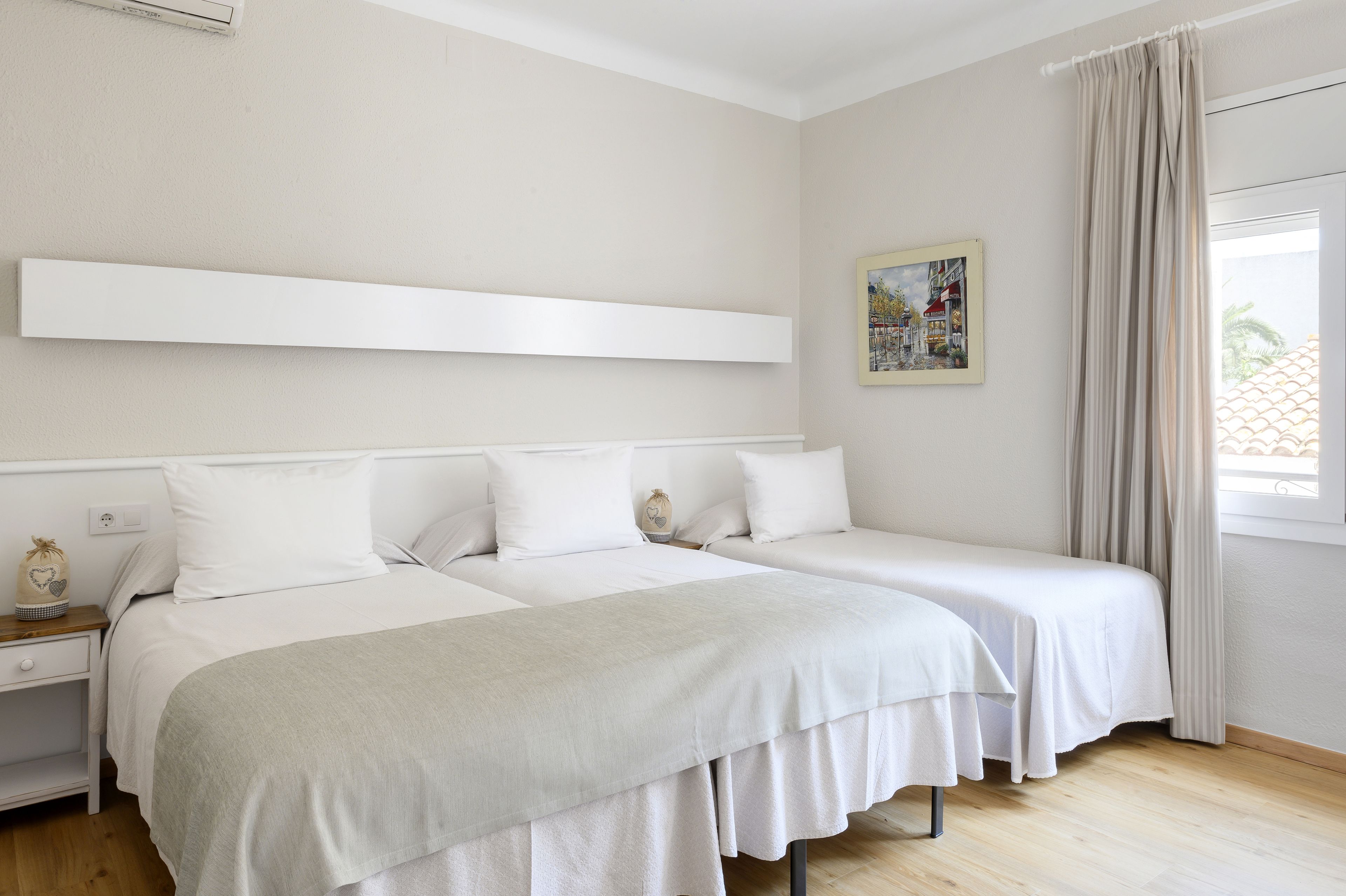 A hotel room with two beds, a white headboard, a bedside table with a lamp, a window with curtains, and a painting on the wall.