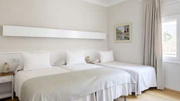 A hotel room with two beds, a white headboard, a bedside table with a lamp, a window with curtains, and a painting on the wall.