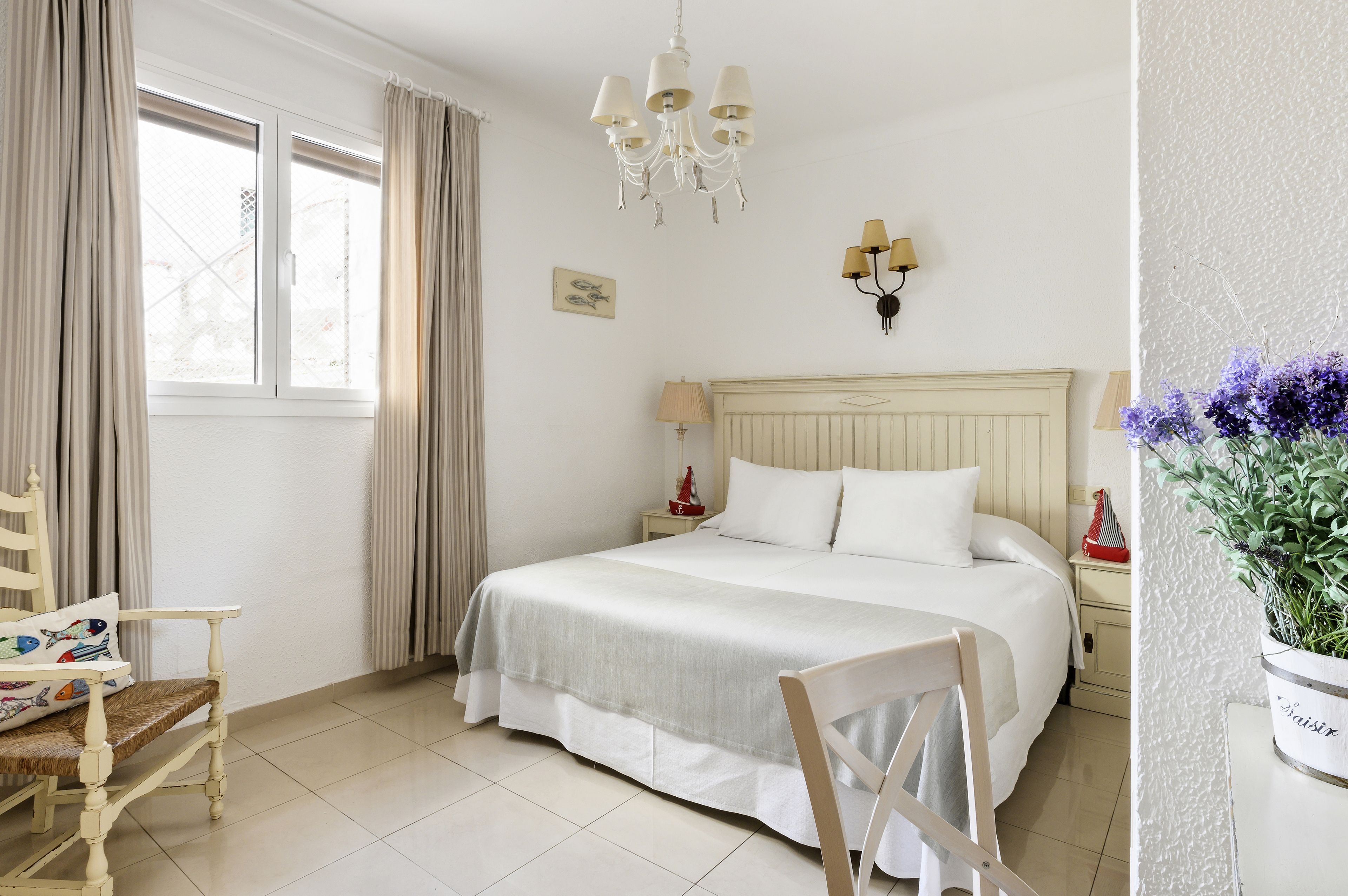 Un dormitorio con cama, una silla, una mesita de noche, una lámpara, un candelabro y una ventana con cortinas.