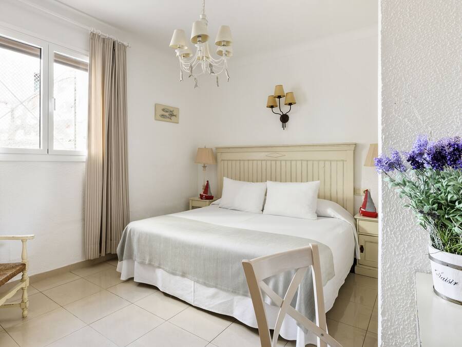 Un dormitorio con cama, una silla, una mesita de noche, una lámpara, un candelabro y una ventana con cortinas.